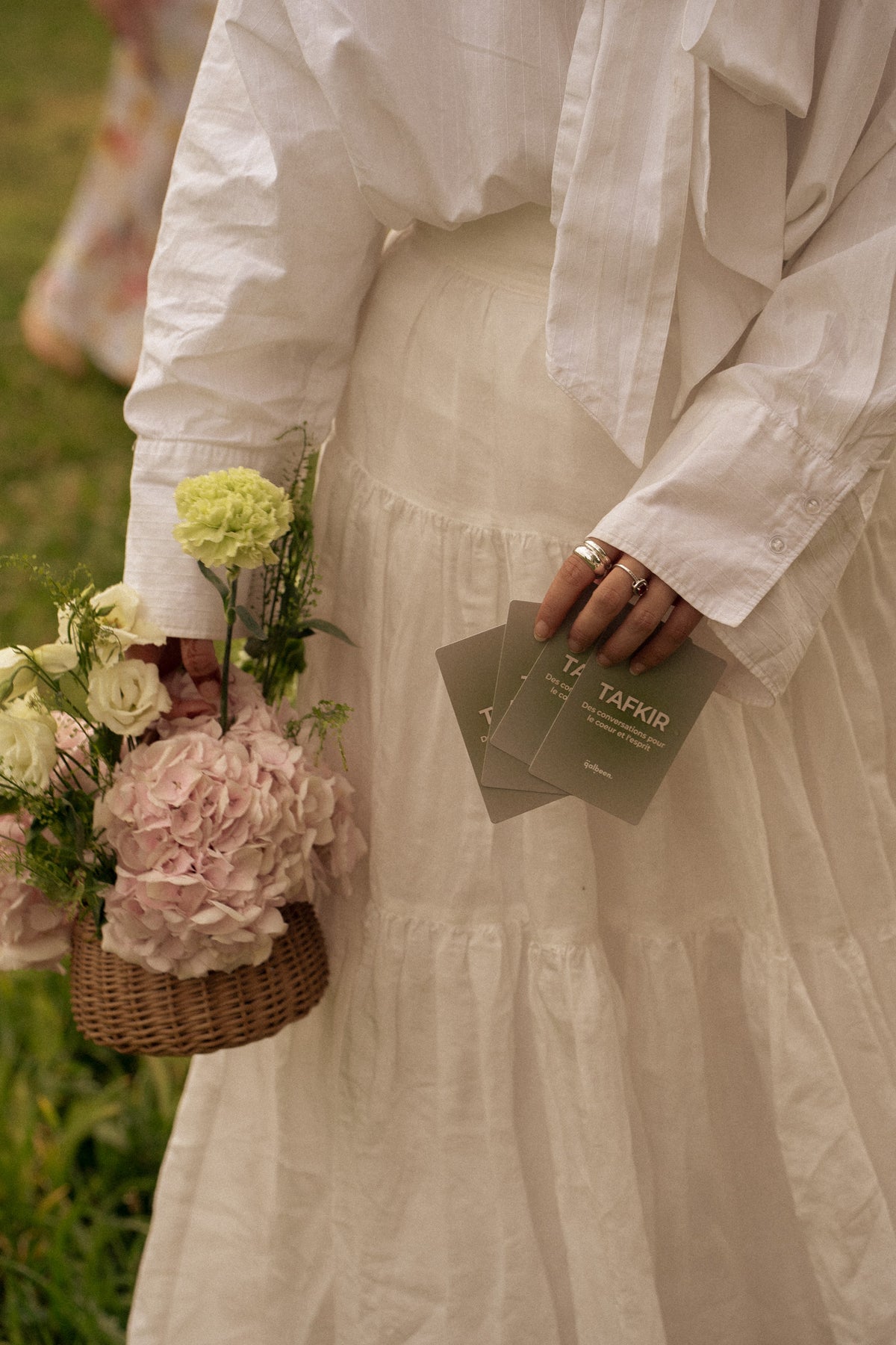 Une femme en robe blanche tient des cartes Qalbeen et un panier de fleurs